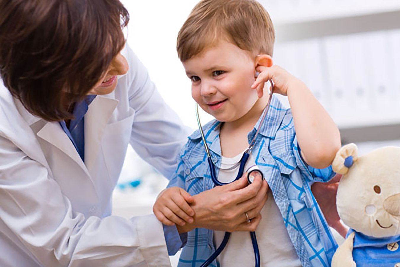 Kinderärztin hört das Herz eines lächelnden kleinen Jungen ab, der spielerisch das Stethoskop hält.